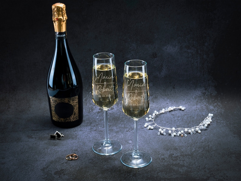 May include: Two champagne flutes with the names Maria and Robert engraved on them, along with the date 09.02.2023. A bottle of champagne and a silver headband with white beads are also in the image.