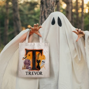 May include: A white canvas tote bag with an orange and black Halloween design. The design features a large letter "T", a spider, a tombstone, a black cat, and a ghost. The bag is personalised with the name "TREVOR".