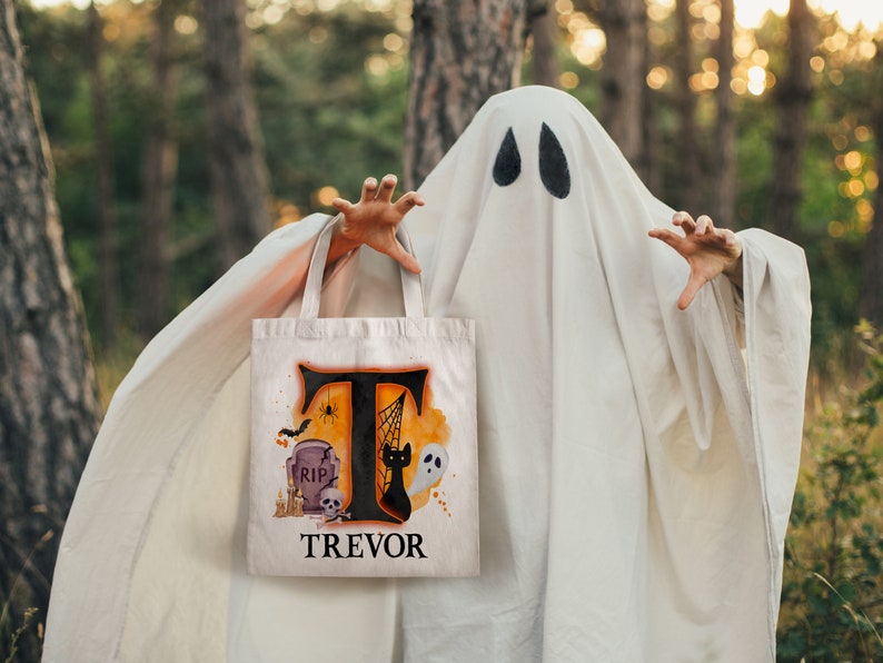 May include: A white canvas tote bag with an orange and black Halloween design. The design features a large letter "T", a spider, a tombstone, a black cat, and a ghost. The bag is personalised with the name "TREVOR".