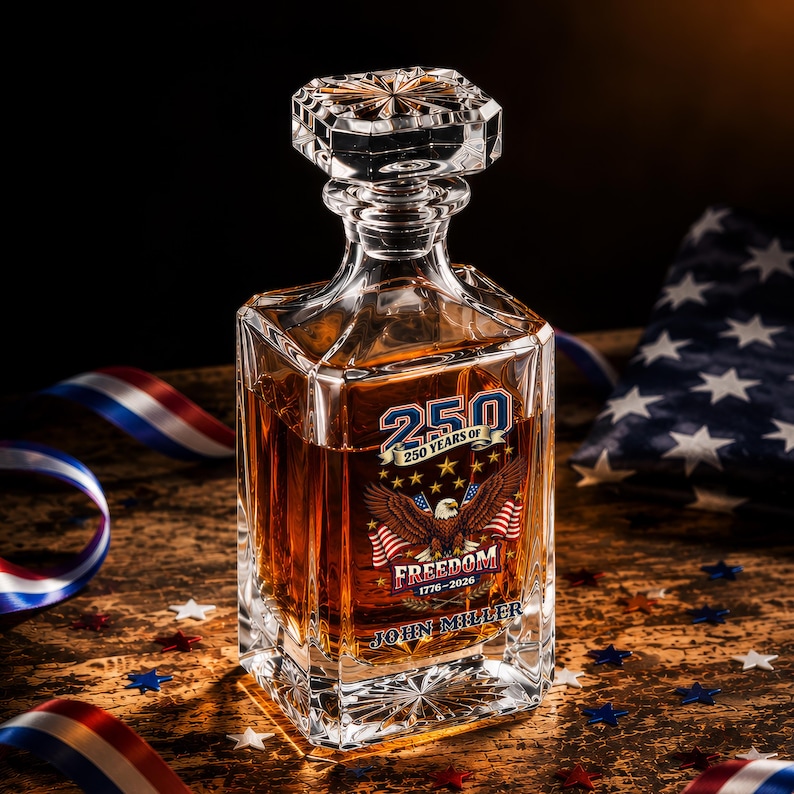 May include: A clear glass decanter filled with amber liquid, featuring a faceted stopper and a square base. The front displays a graphic of a bald eagle, American flags, and the text 250 YEARS OF FREEDOM 1776-2026 and JOHN MILLER.