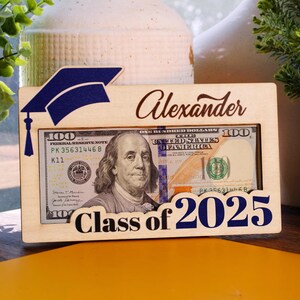 May include: A wooden picture frame with a graduation cap design. The frame has a slot for a photo or money. The frame is personalized with the name "Alexander" and the text "Class of 2026".