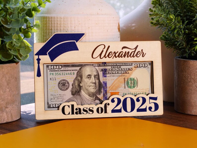 May include: A wooden picture frame with a graduation cap design. The frame has a slot for a photo or money. The frame is personalized with the name "Alexander" and the text "Class of 2026".