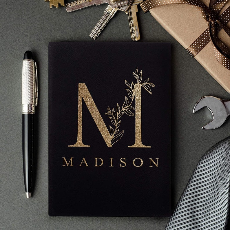Pu&ograve; includere: Un taccuino in pelle nera con un'iniziale "M" in foglia d'oro e il nome "MADISON" stampato sotto. Il taccuino &egrave; circondato da vari oggetti, tra cui chiavi, una chiave inglese e una cravatta.