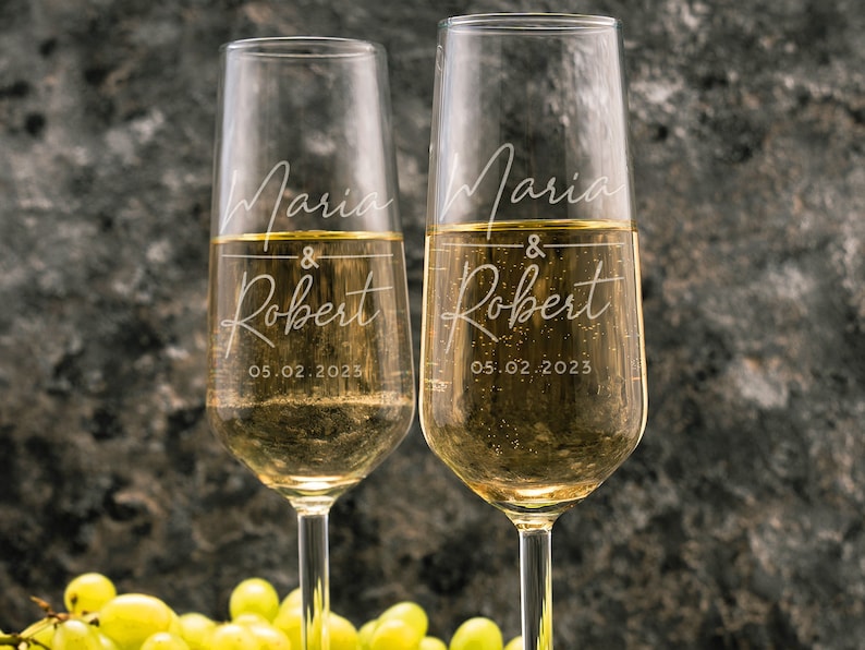 May include: Two champagne flutes with the names Maria & Robert and the date 05.02.2023 etched on them. The flutes are filled with champagne and are set against a textured gray background.