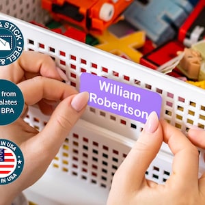 May include: A person is shown applying a purple label that reads "William Robertson" to a white storage bin. The label is peel and stick, and the product is made in the USA. The product is free from phthalates and BPA.