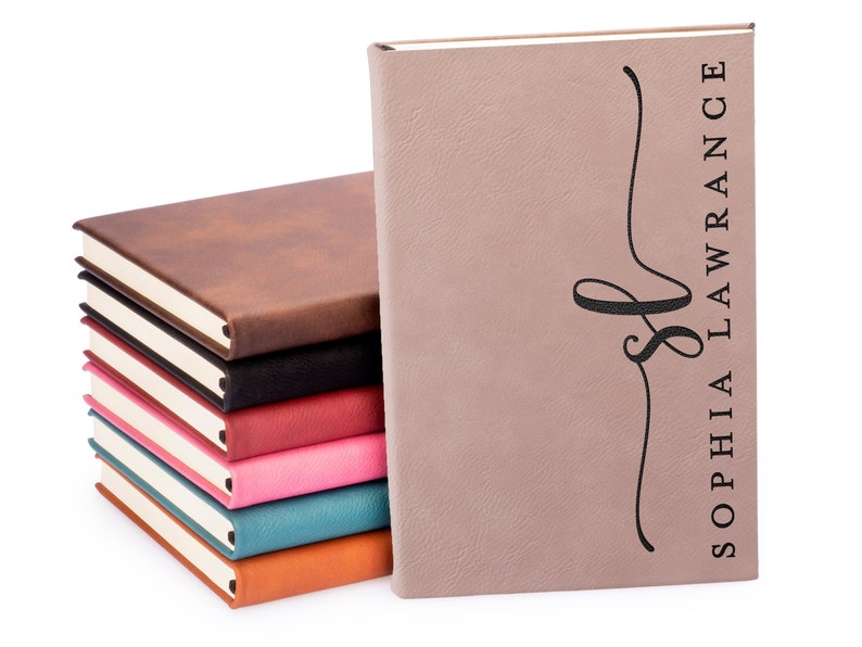 Pu&ograve; includere: Una pila di otto quaderni in similpelle marrone, nero, rosso, rosa, blu e arancione con bordi dorati. Il quaderno in cima &egrave; di colore rosa chiaro con il nome "Sophia Lawrence" e un monogramma "S" stilizzato in nero.