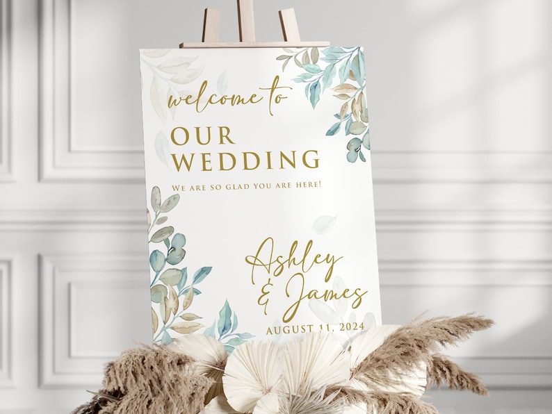 May include: A wedding welcome sign with a white background and gold lettering. The sign features a watercolor design of blue and brown leaves. The text on the sign reads "Welcome to our wedding. We are so glad you are here! Ashley & James August 11, 2024."