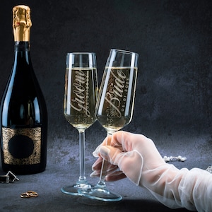 May include: Two champagne flutes with the words "Groom" and "Bride 06.01.2023" etched on them. A bottle of champagne is in the background.