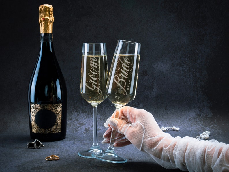 May include: Two champagne flutes with the words "Groom" and "Bride 06.01.2023" etched on them. A bottle of champagne is in the background.