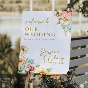 May include: A white wedding sign with gold lettering and watercolor floral designs. The sign reads "Welcome to Our Wedding, We are so glad you are here! Jessica & Chris, October 21, 2024".