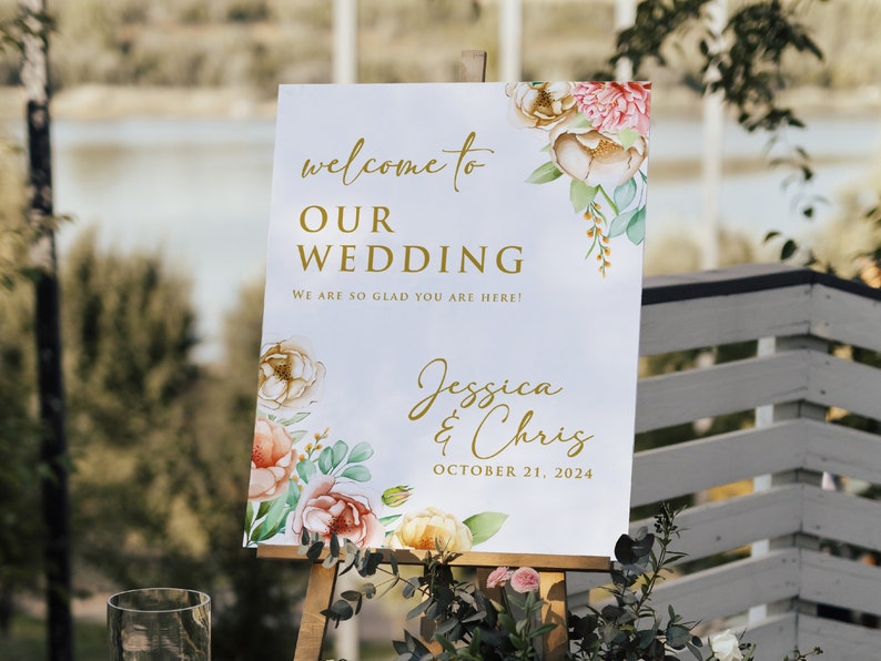 May include: A white wedding sign with gold lettering and watercolor floral designs. The sign reads "Welcome to Our Wedding, We are so glad you are here! Jessica & Chris, October 21, 2024".