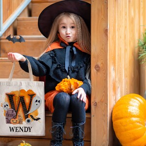 May include: A child dressed as a witch is holding a white canvas tote bag with a black letter W, a ghost, a tombstone, a pumpkin, a spider web, and an eye. The bag says "WENDY" at the bottom. The child is sitting on wooden steps with a pumpkin in their lap.