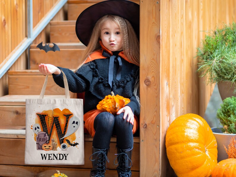 May include: A child dressed as a witch is holding a white canvas tote bag with a black letter W, a ghost, a tombstone, a pumpkin, a spider web, and an eye. The bag says "WENDY" at the bottom. The child is sitting on wooden steps with a pumpkin in their lap.