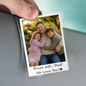May include: A clear plastic photo holder with a family photo. The photo shows a man hugging two children. Text at the bottom reads "Drive safe, Dad! We Love You!" with a heart symbol. The holder is held by a hand.