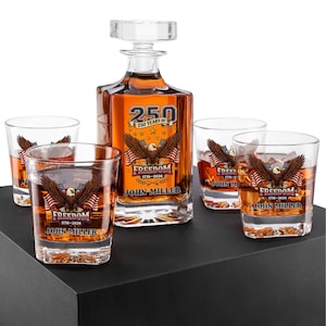 May include: A clear glass decanter with a square stopper and four matching whiskey glasses. Each piece features a graphic of a bald eagle, American flags, and the text 250 YEARS OF FREEDOM 1776-2026 JOHN MILLER printed in blue, red, and gold.