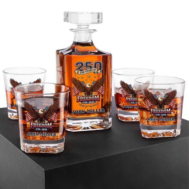 May include: A clear glass decanter with a square stopper and four matching whiskey glasses. Each piece features a graphic of a bald eagle, American flags, and the text 250 YEARS OF FREEDOM 1776-2026 JOHN MILLER printed in blue, red, and gold.