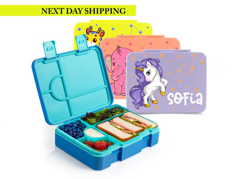 May include: A blue and turquoise lunchbox with compartments, filled with food. Several closed lunchboxes with animal and unicorn designs are in the background. The text "SOFIA" is on one lunchbox.