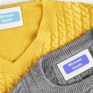 May include: Close-up of two sweaters, one yellow and one gray, with labels that read "Michael Smith." The yellow sweater has a cable knit design, while the gray sweater has a simpler knit. The labels are light blue with white text.