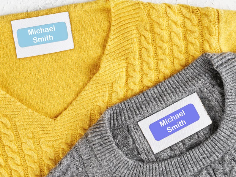 May include: Close-up of two sweaters, one yellow and one gray, with labels that read "Michael Smith." The yellow sweater has a cable knit design, while the gray sweater has a simpler knit. The labels are light blue with white text.