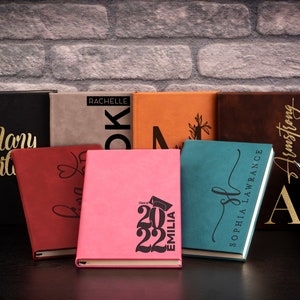 Pu&ograve; includere: Una collezione di otto diari personalizzati con lettere dorate su copertine di diversi colori. I diari sono disposti in fila su una superficie nera. I diari hanno nomi, iniziali e l'anno 2022 stampati su di essi.