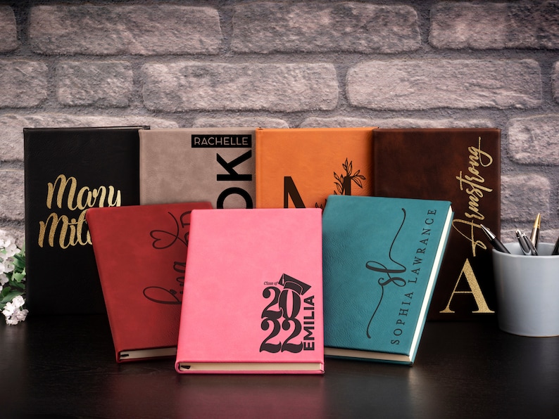 Pu&ograve; includere: Una collezione di otto diari personalizzati con lettere dorate su copertine di diversi colori. I diari sono disposti in fila su una superficie nera. I diari hanno nomi, iniziali e l'anno 2022 stampati su di essi.