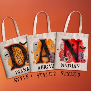 May include: Three Halloween-themed tote bags with different designs. The first bag features a black letter "D" with a spiderweb and a ghost, and the name "Diana" below. The second bag features a black letter "A" with a ghost, an eye, and a bone, and the name "Abigail" below. The third bag features a black letter "N" with a ghost, a bat, and a pumpkin, and the name "Nathan" below. Each bag has the words "Style 1", "Style 2", and "Style 3" below the name.