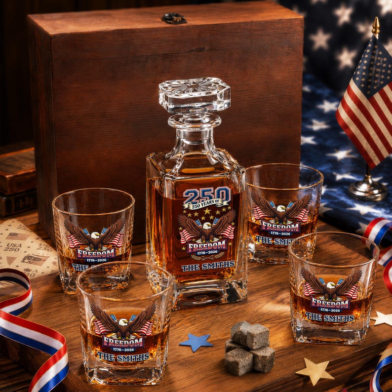 May include: A glass decanter and four matching whiskey glasses filled with amber liquid, featuring a bald eagle graphic, American flags, and the text 250 YEARS OF FREEDOM 1776-2026 THE SMITHS. Includes a wooden box and stone whiskey cubes.