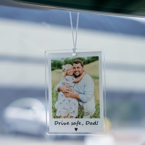May include: Clear acrylic car air freshener with a photo of a man and a child. The photo has the text "Drive safe, Dad!" and a heart symbol. The air freshener hangs from a string, and is hanging from a car's rearview mirror.