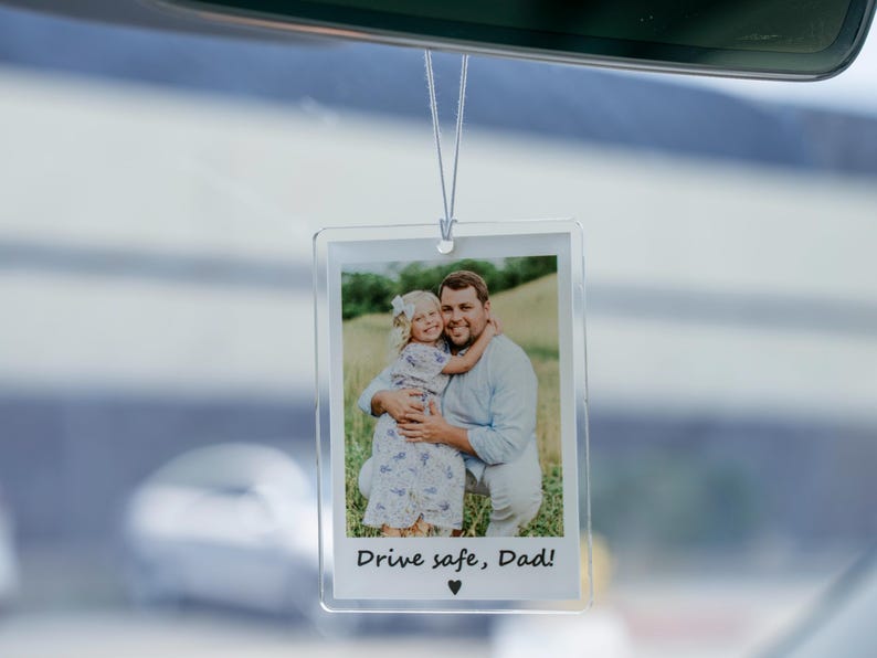 May include: Clear acrylic car air freshener with a photo of a man and a child. The photo has the text "Drive safe, Dad!" and a heart symbol. The air freshener hangs from a string, and is hanging from a car's rearview mirror.