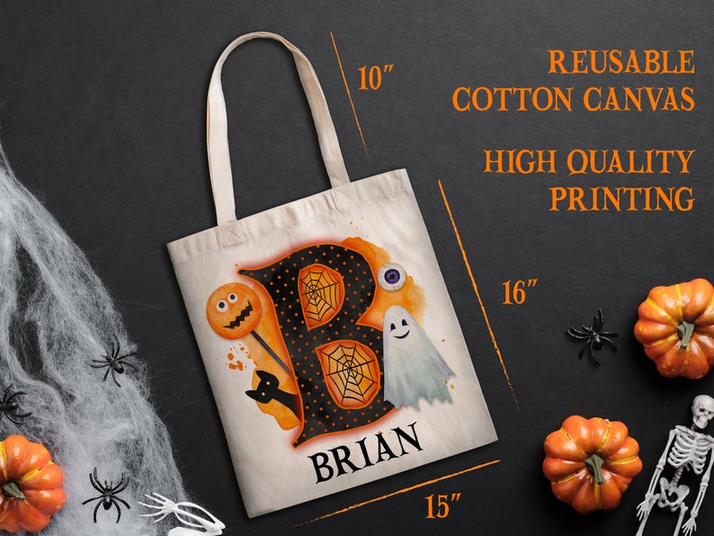 May include: A reusable cotton canvas tote bag with a black and orange Halloween-themed design featuring the letter "B" with a spiderweb, a ghost, a lollipop with a jack-o-lantern face, and a black cat. The bag is personalised with the name "BRIAN" in black lettering. The bag measures 40.6 cm by 38.1 cm.
