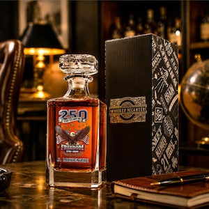 May include: A square glass whiskey decanter with a faceted stopper, filled with amber liquid. The front features a graphic with a bald eagle, American flags, and the text 250 YEARS OF FREEDOM 1776-2026 and J. MCKENZIE. Beside it stands a black box.