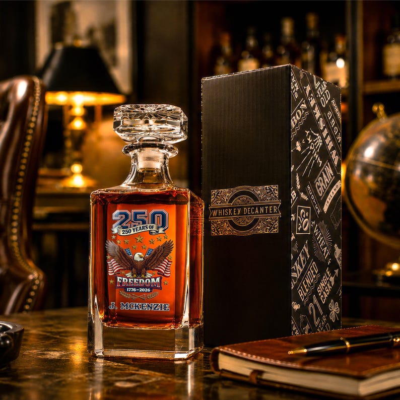 May include: A square glass whiskey decanter with a faceted stopper, filled with amber liquid. The front features a graphic with a bald eagle, American flags, and the text 250 YEARS OF FREEDOM 1776-2026 and J. MCKENZIE. Beside it stands a black box.