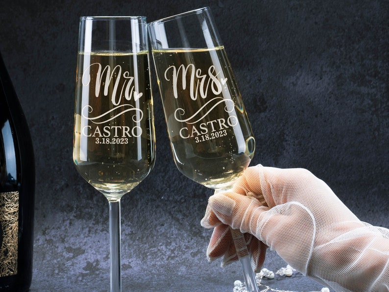 May include: Two champagne flutes with the words "Mr. Castro 3.18.2023" and "Mrs. Castro 3.18.2023" etched on them. The flutes are filled with champagne and are being held by a hand wearing a white glove.