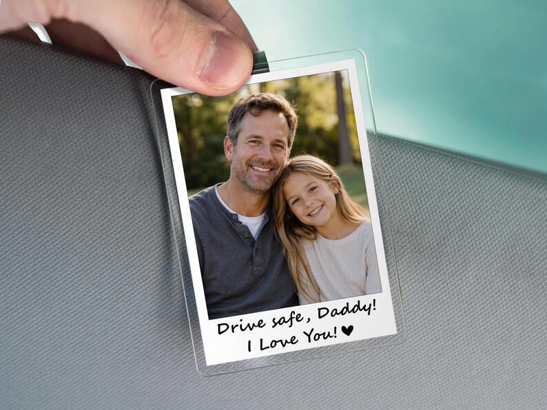 May include: Clear plastic photo holder with a picture of a man and a girl. The photo has the text "Drive safe, Daddy! I Love You!" in black. The holder is being held by a hand.