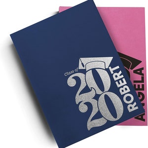 Pu&ograve; includere: Un biglietto di laurea blu con lettere argentate glitter che dicono "Classe del 2020" e un cappello di laurea. Il biglietto &egrave; piegato a met&agrave; e ha un biglietto rosa sopra con lettere nere che dicono "Angela Robert".