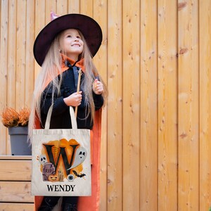 May include: A white canvas tote bag with an orange and black Halloween design. The bag features a large letter "W" with a ghost, a pumpkin, a tombstone, and an eye. The text "WENDY" is printed below the design.