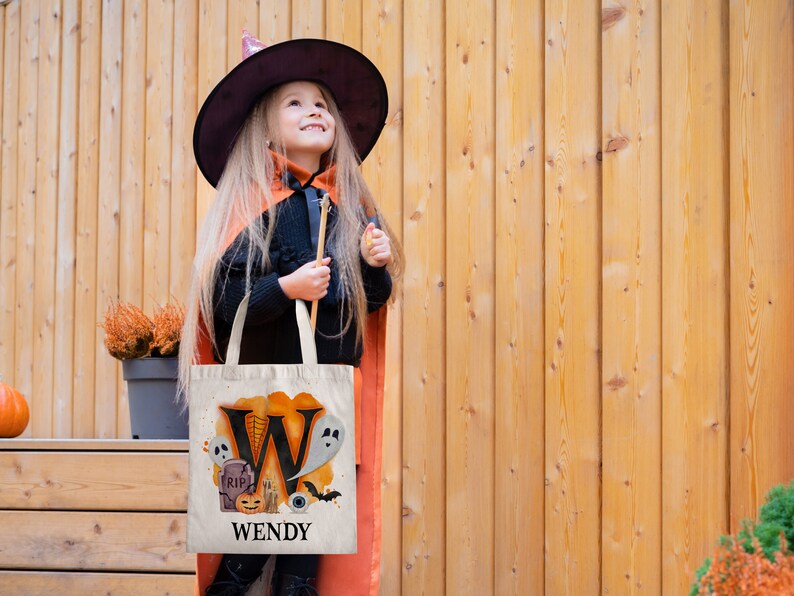 May include: A white canvas tote bag with an orange and black Halloween design. The bag features a large letter "W" with a ghost, a pumpkin, a tombstone, and an eye. The text "WENDY" is printed below the design.