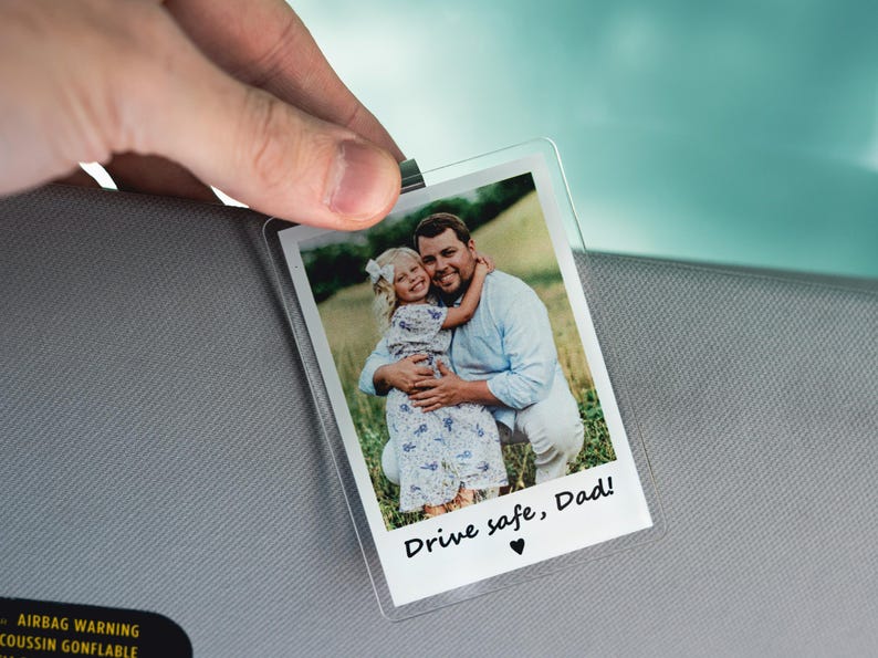 May include: Clear plastic photo holder with a picture of a man and a young girl hugging. The photo has the text "Drive safe, Dad!" and a heart symbol. The holder is being held up.