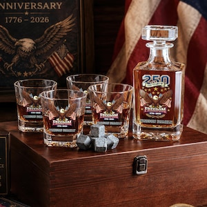 May include: A glass decanter and three matching whiskey glasses filled with amber liquid, each featuring a bald eagle graphic, the text 250 YEARS OF FREEDOM 1776-2026, and WATSON FAMILY. A small pile of grey stone whiskey cubes sits in front of the glasses.