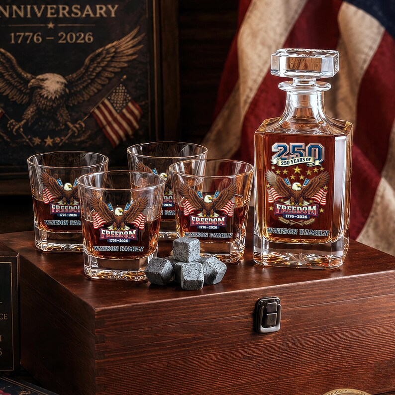 May include: A glass decanter and three matching whiskey glasses filled with amber liquid, each featuring a bald eagle graphic, the text 250 YEARS OF FREEDOM 1776-2026, and WATSON FAMILY. A small pile of grey stone whiskey cubes sits in front of the glasses.