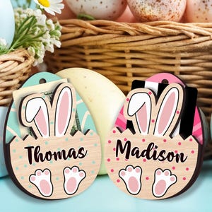May include: Wooden Easter egg-shaped gift card holders with bunny ears and paw prints. One holder is light blue with the name "Thomas", the other is pink with the name "Madison". Both have a slot for a gift card or money.