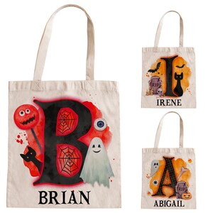 May include: Three Halloween-themed tote bags with personalised names. The bags are made of natural canvas and feature a black letter with a watercolour background and Halloween-themed illustrations. The bags are labelled "BRIAN", "IRENE", and "ABIGAIL".