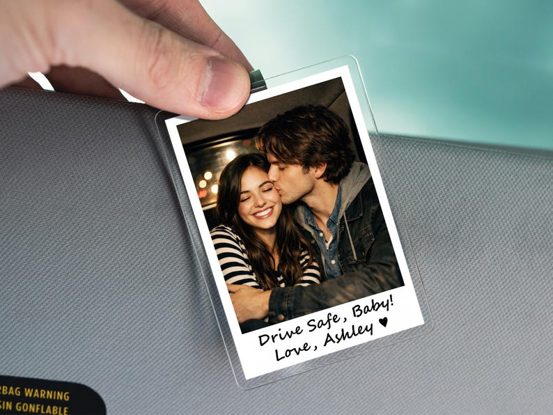 May include: A clear plastic photo holder with a picture of a couple kissing. The photo has the text "Drive Safe, Baby! Love, Ashley" in black script. The holder is clipped to a gray surface.