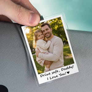 May include: A clear, rectangular photo holder with a picture of a man and a child. The photo has the text "Drive safe, Daddy! I Love You!" in black. The holder is clipped to a gray car visor.