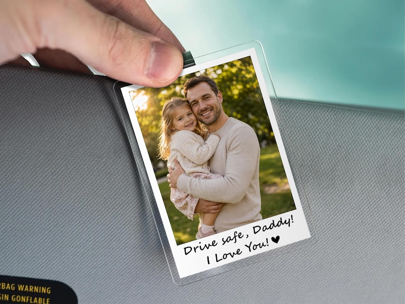 May include: A clear, rectangular photo holder with a picture of a man and a child. The photo has the text "Drive safe, Daddy! I Love You!" in black. The holder is clipped to a gray car visor.
