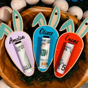 May include: Wooden carrot-shaped Easter gift holders with bunny ears, each holding rolled-up money. The carrots are in pastel colors: lavender, blue, and orange, with names "Amelia", "Oliver", and "Lucas". The holders are in a wooden bowl.