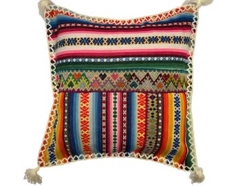May include: A colorful throw pillow with a geometric pattern in shades of red, orange, yellow, green, blue, and pink. The pillow has a white fringe border and tassels on the corners.