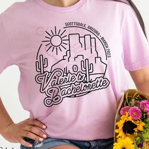 Op de afbeelding: Een roze T-shirt met een zwart grafisch ontwerp met een woestijnlandschap, cactussen en de tekst "Valerie's Bachelorette" met de locatie "Scottsdale, Arizona - maart 2023".