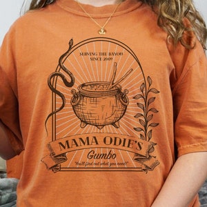 May include: A woman wearing an orange t-shirt with a vintage-style illustration of a cauldron and the text "Mama Odie's Gumbo" and "Serving the Bayou Since 2009".