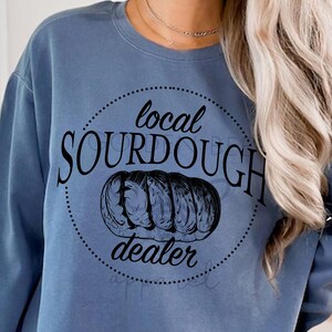 Può includere: Felpa blu jeans con grafica circolare nera con la scritta "local SOURDOUGH dealer". La grafica include un'illustrazione di pane. Le parole "BLUE JEAN" sono stampate in nero in basso a destra.